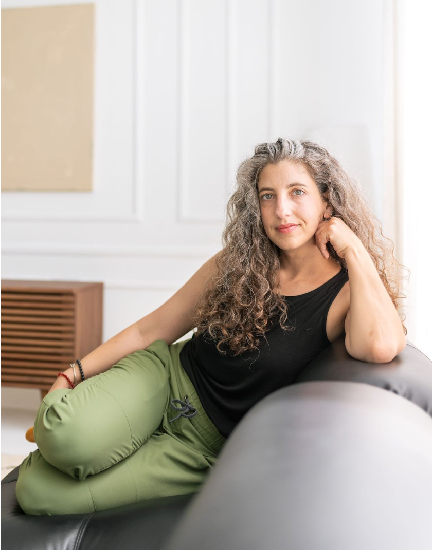 Image of Emily with long curly hair, sitting with head propping up arm comfortably on a grey sofa
